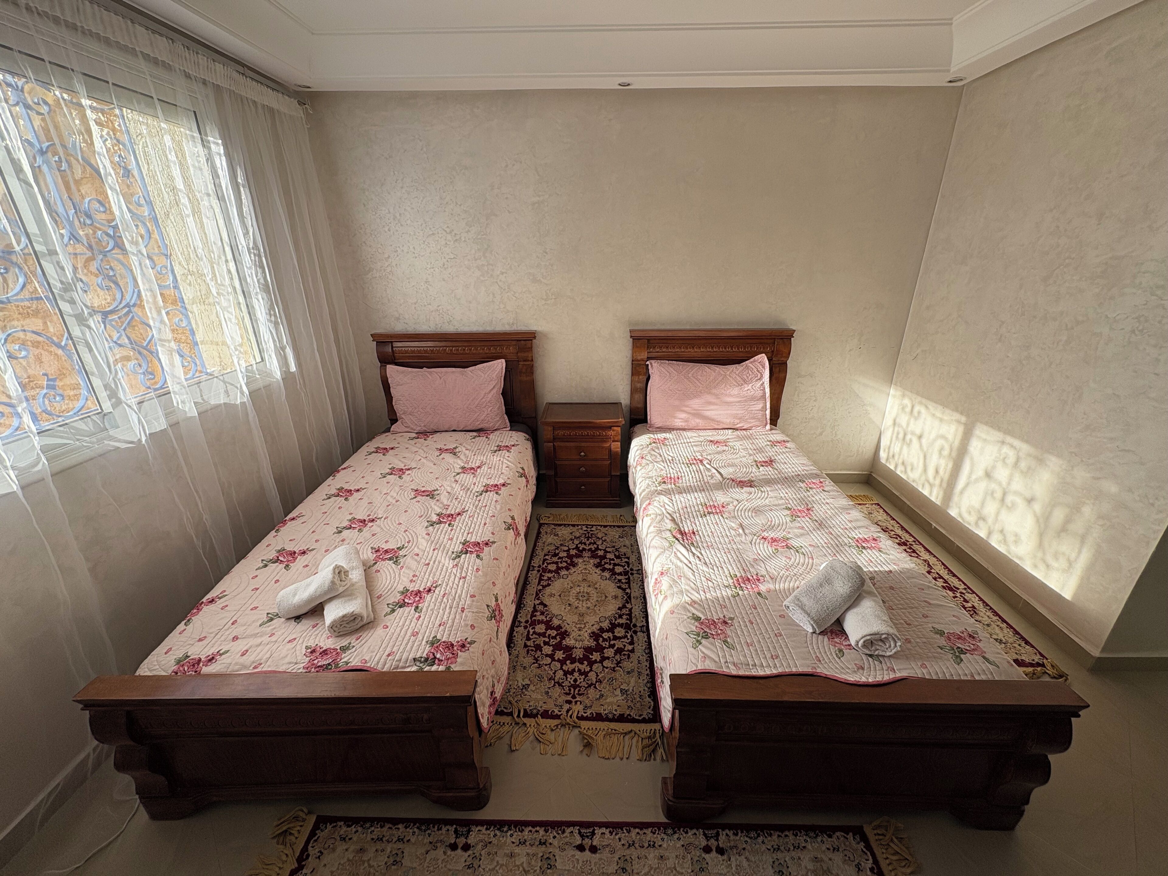Une chambre double avec deux lits simples, une table de chevet en bois, un tapis à motifs et une fenêtre avec des rideaux.