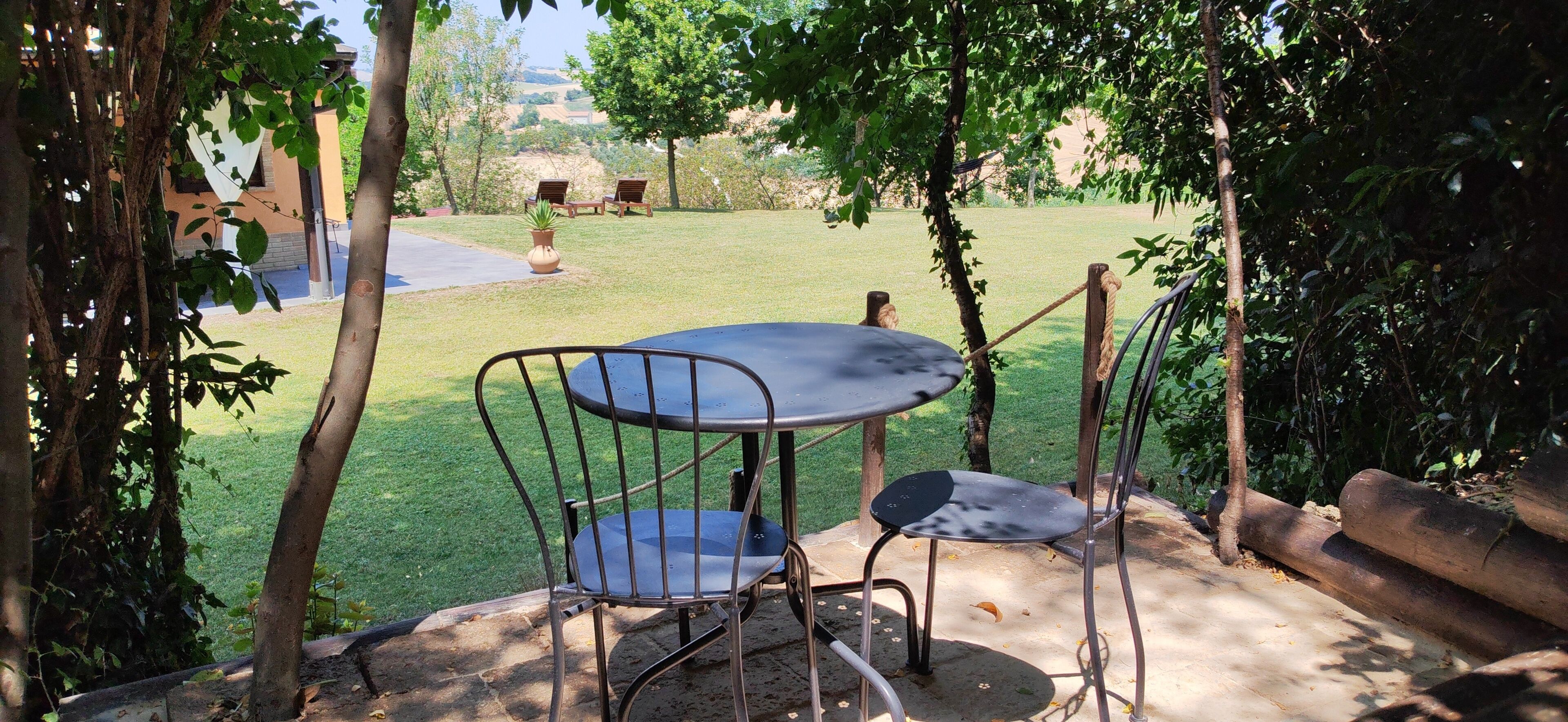 An outdoor seating area with a round table and two chairs, surrounded by trees and a grassy lawn.