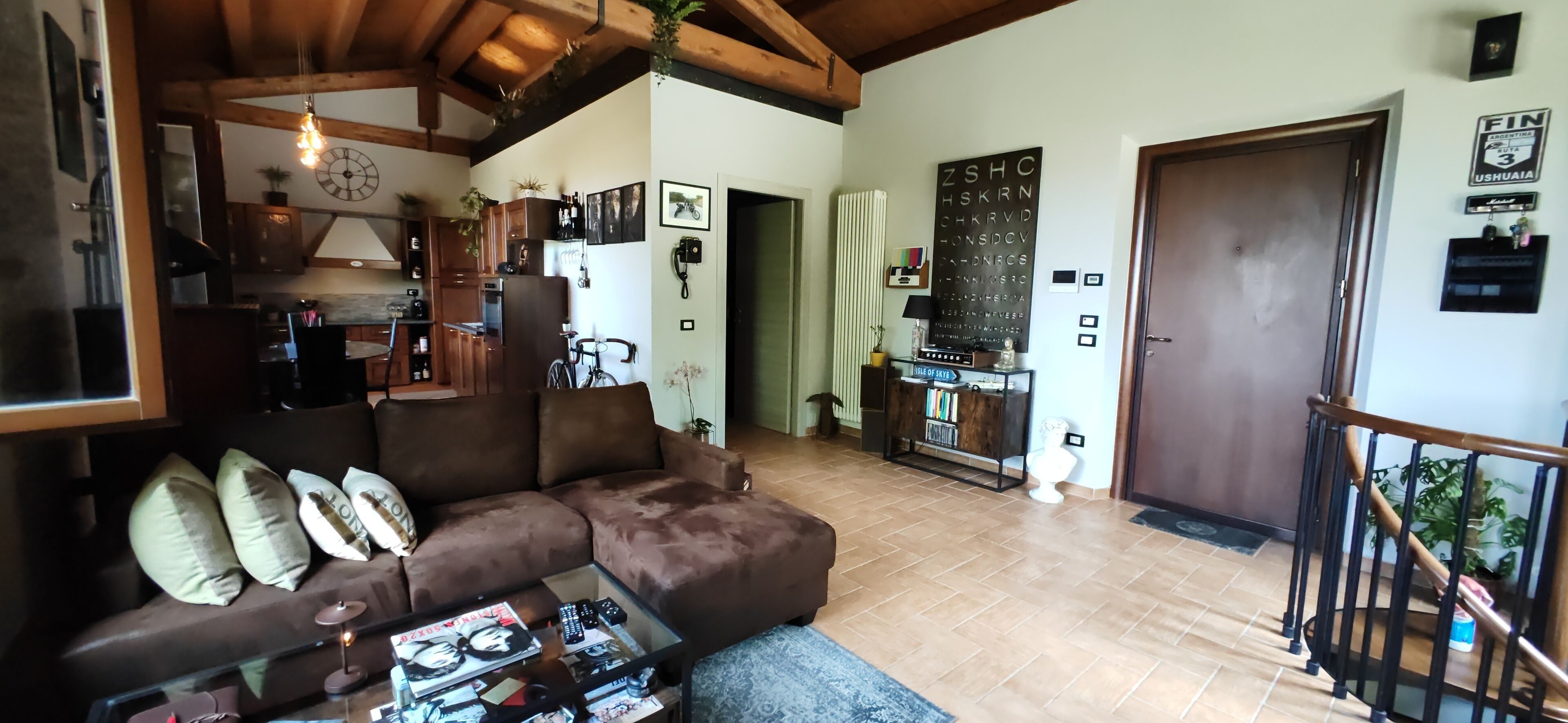 A cozy living room with a brown sectional sofa, a glass coffee table, and a kitchen area with wooden cabinets and a refrigerator.