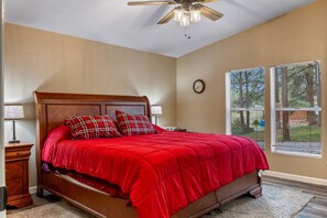 A bedroom with a wooden bed, red bedding, a ceiling fan, a clock, and windows with blinds.