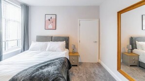 Ein Schlafzimmer mit Bett, Nachttischen, einem Spiegel, einer Tür und einem Fenster mit Vorhängen.