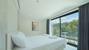 Une chambre moderne avec un grand lit, une vue sur la verdure et un balcon aménagé avec du mobilier d’extérieur.