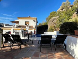 A pool area with black lounge chairs, a white building with a tiled roof, and a view of a mountain in the background. - Spanish country house. Completely free, yet two restaurants within walking distance. (COMARES)