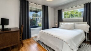 A bedroom with a bed, a desk, and a chair. There are two windows with curtains, one on each side of the bed. - Division House Extended Stay (Walla Walla)