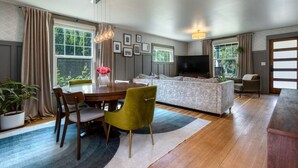 A modern living room with a sofa, TV, dining table, chairs, and a large window with curtains. - Division House Extended Stay (Walla Walla)