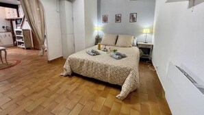 Quarto com piso de madeira, uma cama com roupa de cama estampada, uma mesinha de cabeceira com abajur e um guarda-roupa.