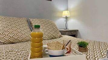 Cama bem arrumada com roupa de cama estampada, bandeja com itens de café da manhã, garrafa de suco de laranja e uma pequena planta na mesa de cabeceira.
