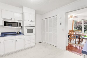 A kitchen with white cabinets, a microwave, an oven, and a sink. There is a dining area with a table and chairs.
