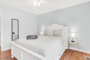 A bedroom with a bed, a bedside table with a lamp, a mirror, and a wooden floor.