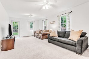 A spacious living room with a large black leather sofa, a brown leather sofa, and a brown armchair. There is a flat-screen TV on a wooden stand, a ceiling fan, and a large window with curtains.
