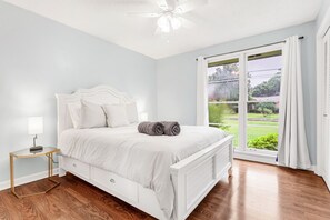 Een slaapkamer met een wit bed, een nachtkastje met een lamp, en uitzicht op een groen landschap door een groot raam.