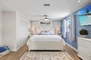 Une chambre avec un grand lit, un ventilateur de plafond, une table de chevet avec des lampes et une vue sur l’océan par une fenêtre.