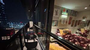 A balcony with a dining table and chairs, a bed with a red and yellow rug, and a city view at night.