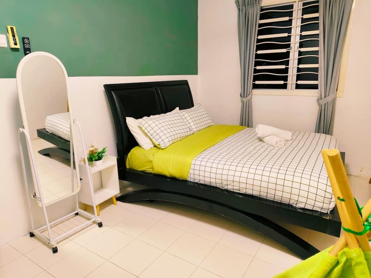 A bed with a black frame, a white headboard, and a green blanket. There is a white bedside table with a plant on it. A white wardrobe is visible in the background.