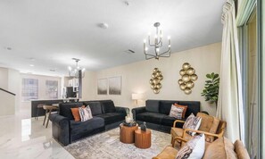 Ein modernes Wohnzimmer mit schwarzen Sofas, einem Holztisch und einer Kristallleuchter. Im Hintergrund ist ein Essbereich mit einer sichtbaren Küche zu sehen.