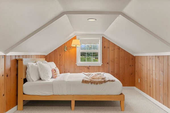 A bedroom with a sloped ceiling, wooden panel walls, a bed with white bedding, a window with a view, and a wall-mounted lamp.