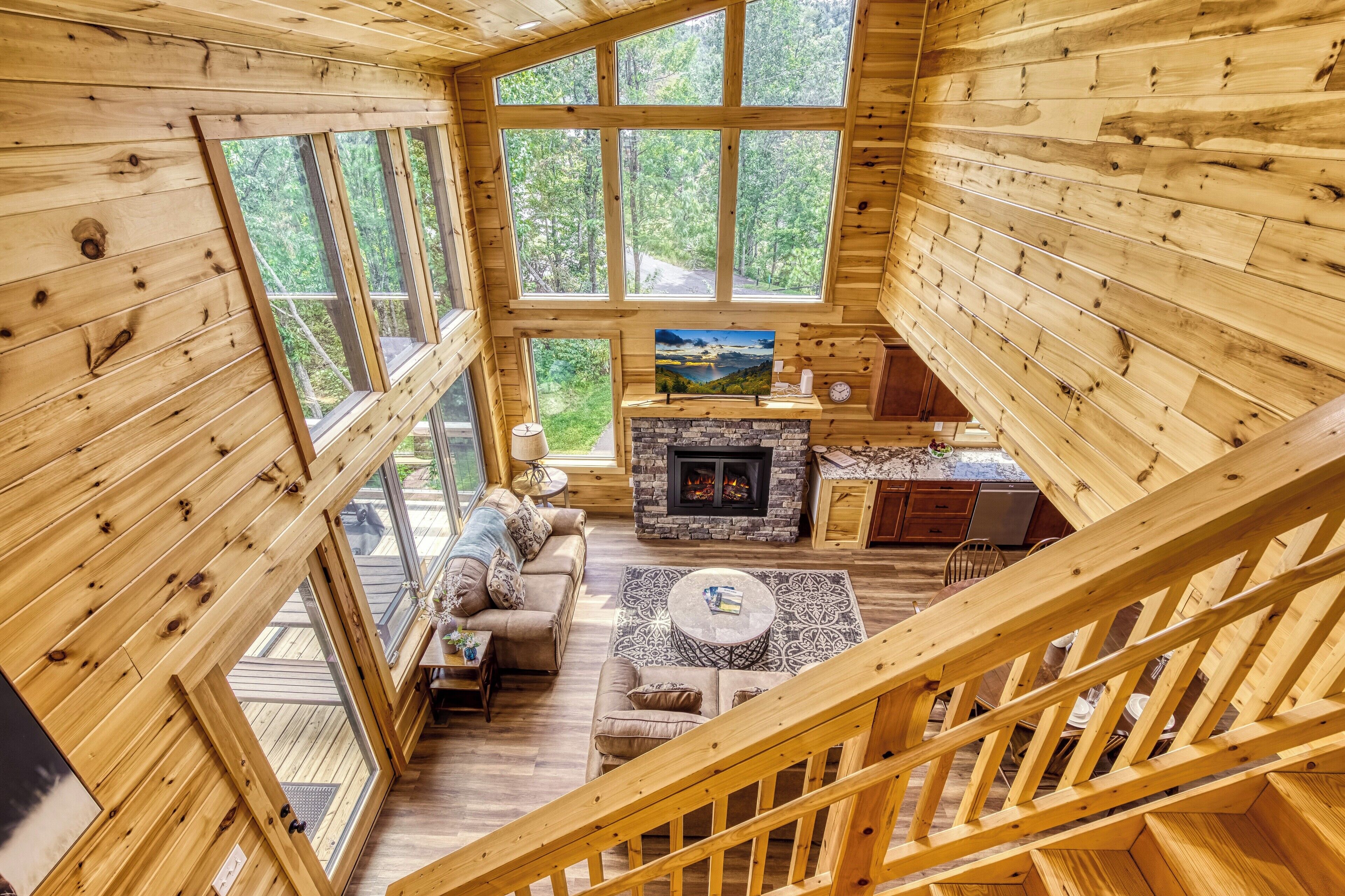 A wooden cabin interior with a loft bedroom, a fireplace, a sofa set, and large windows offering a view of the forest.
