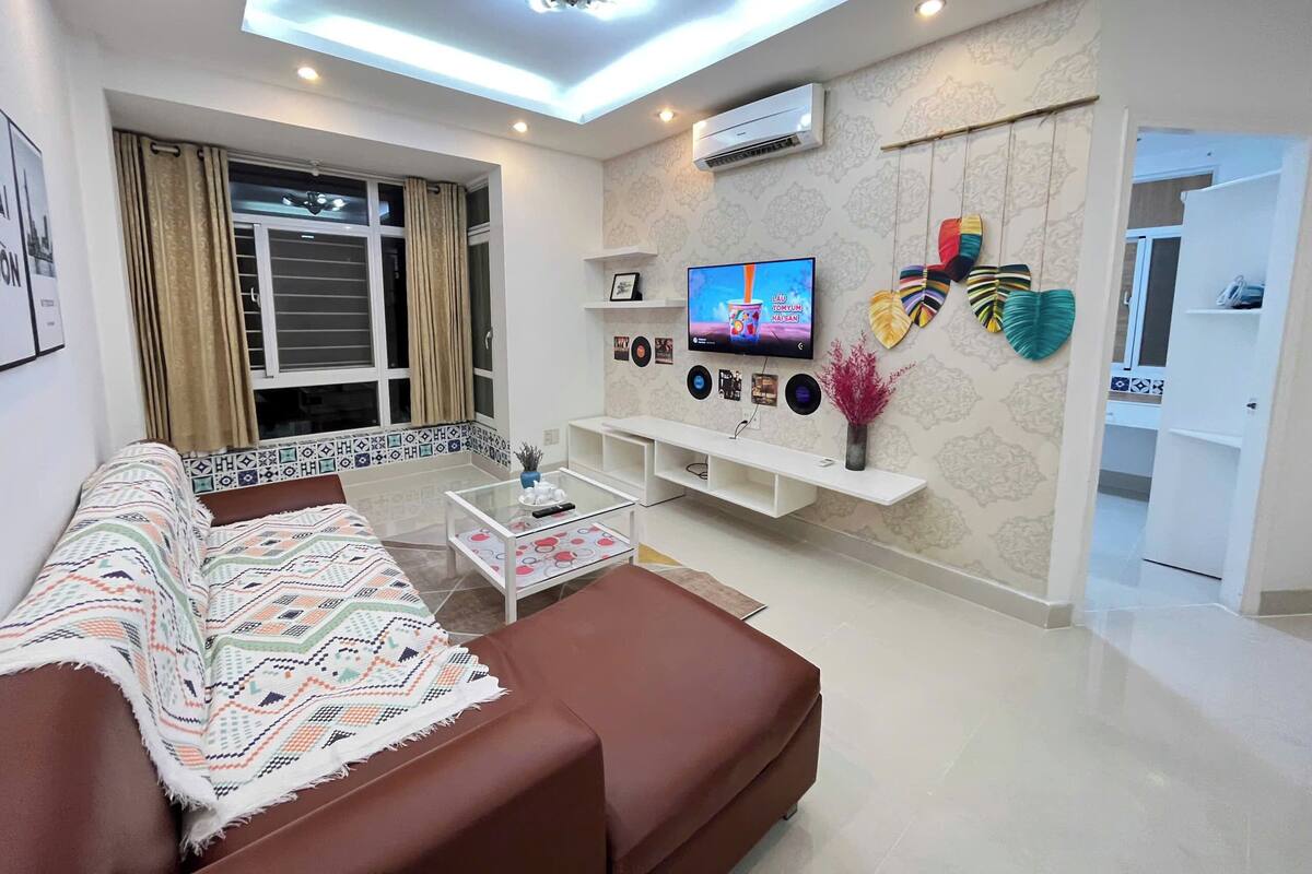 Een moderne woonkamer met een bruine bank, een glazen salontafel, een aan de muur bevestigde televisie en decoratieve objecten.