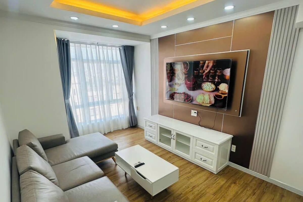 Een moderne woonkamer met een hoekbank, een witte salontafel en een televisie die aan een wand is bevestigd, met een houten kast eronder.