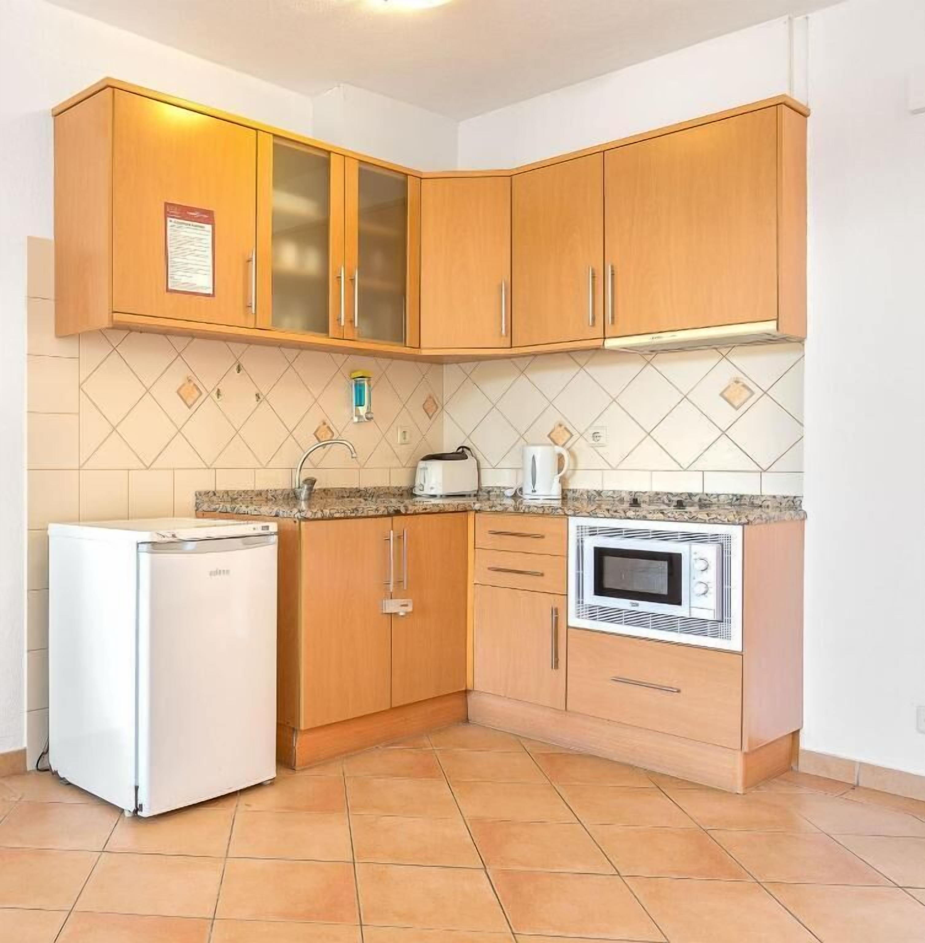 Eine kleine Küche mit Holzausstattung, einem weißen Kühlschrank, einer Mikrowelle und einem Wasserkocher auf der Arbeitsfläche.