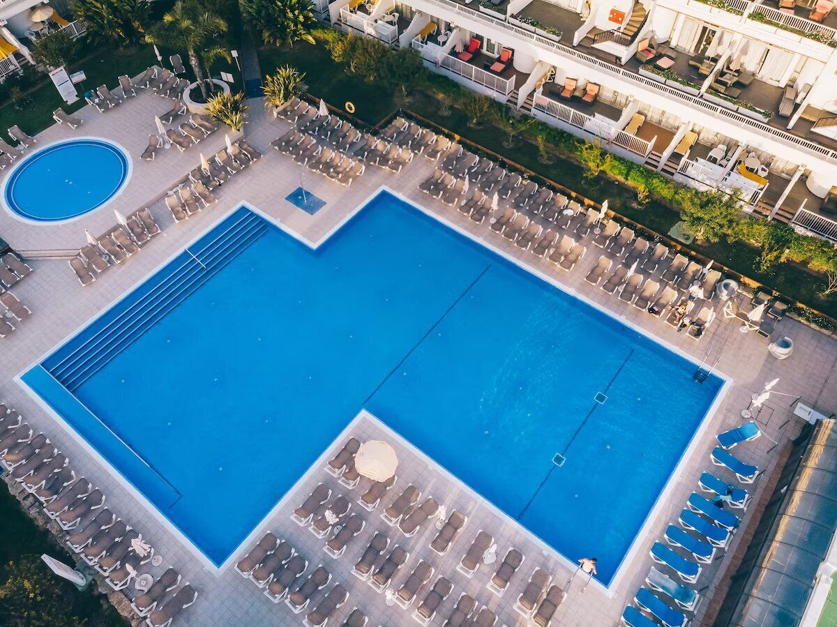 Luftaufnahme eines Hotel-Poolbereichs mit mehreren Liegestühlen und einem zentralen Schwimmbecken.