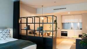 Una habitación de hotel moderna con cama, escritorio, kitchenette y una pared de vidrio.