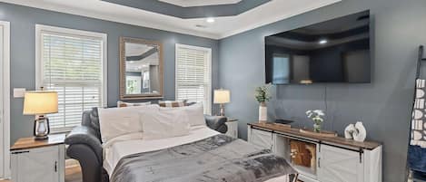 A bedroom with a bed, bedside tables, a dresser, a mirror, a TV, and a window with blinds.