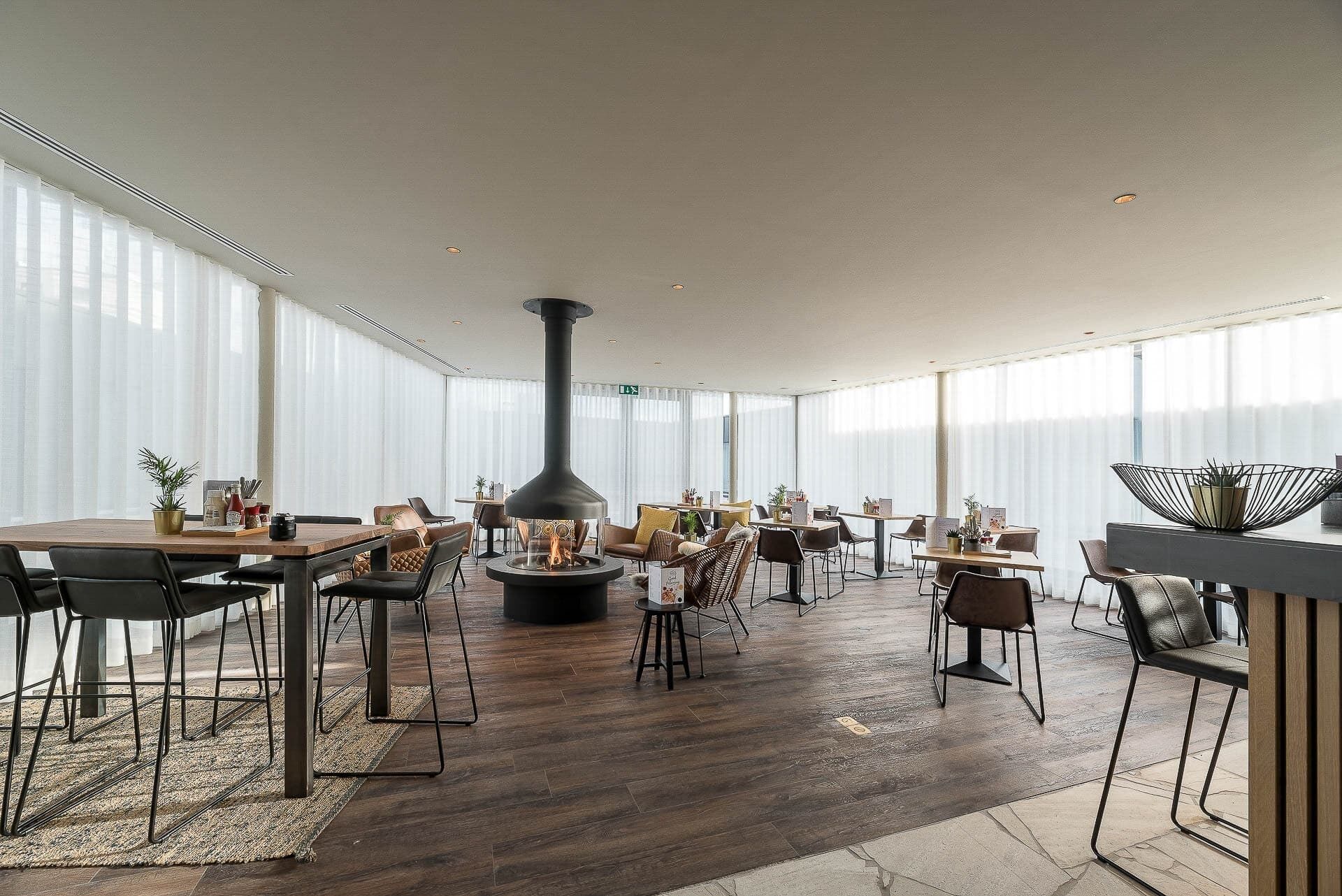 A spacious indoor area with multiple tables and chairs, a central fireplace, and large windows allowing natural light.