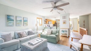A modern living room with a sofa, ottoman, and a dining area. - Duck Condo, 5 min walk to beach w/ community pool! (Duck)