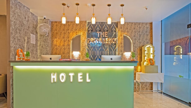 A hotel reception desk with a green counter, two laptops, a fan, and a decorative wall with the text "THE PROMINENCE". - The Prominence Hotel Restaurant & Bar (Greater Noida)