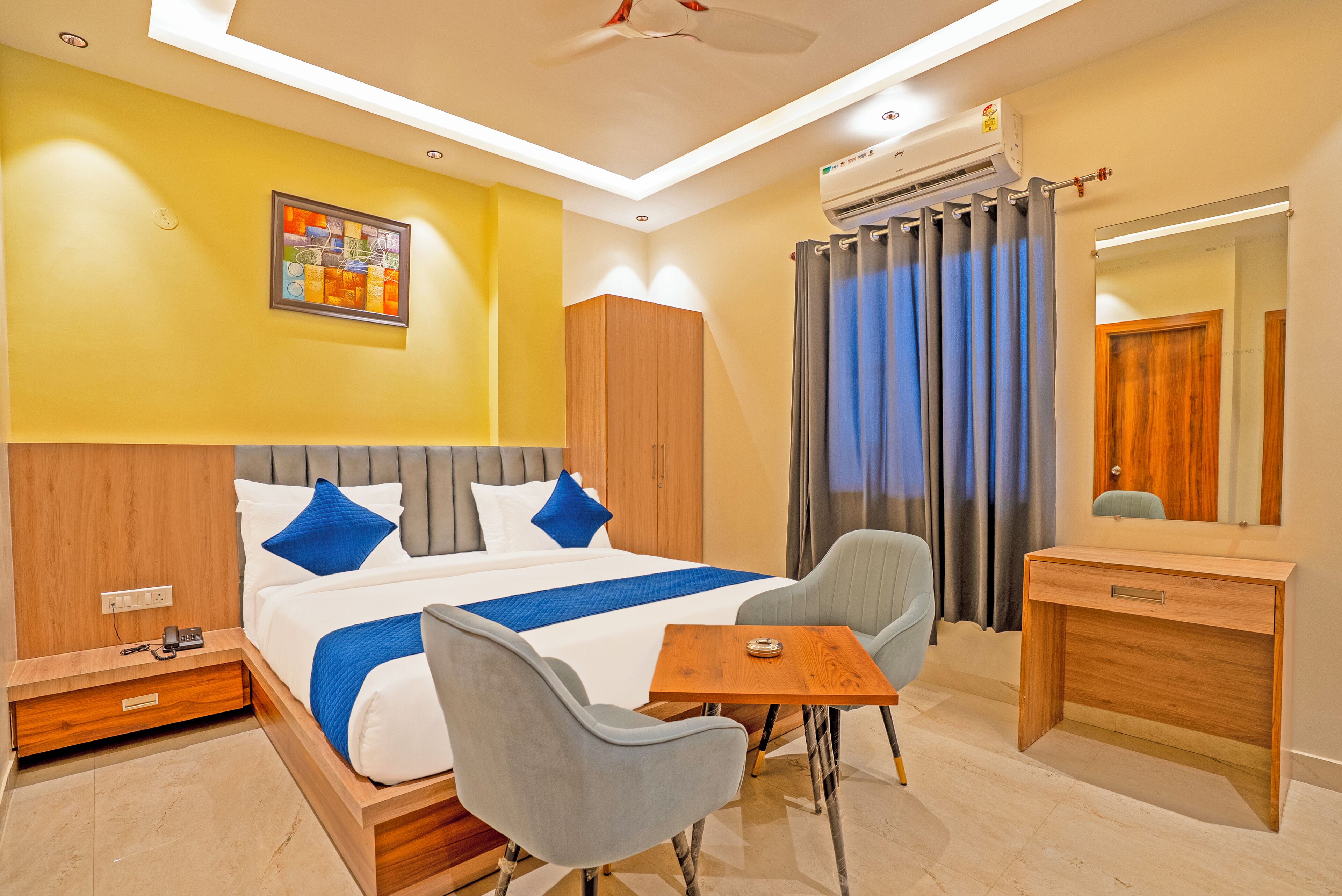 A hotel room with a bed, a desk, a chair, a bedside table, a mirror, and an air conditioning unit.