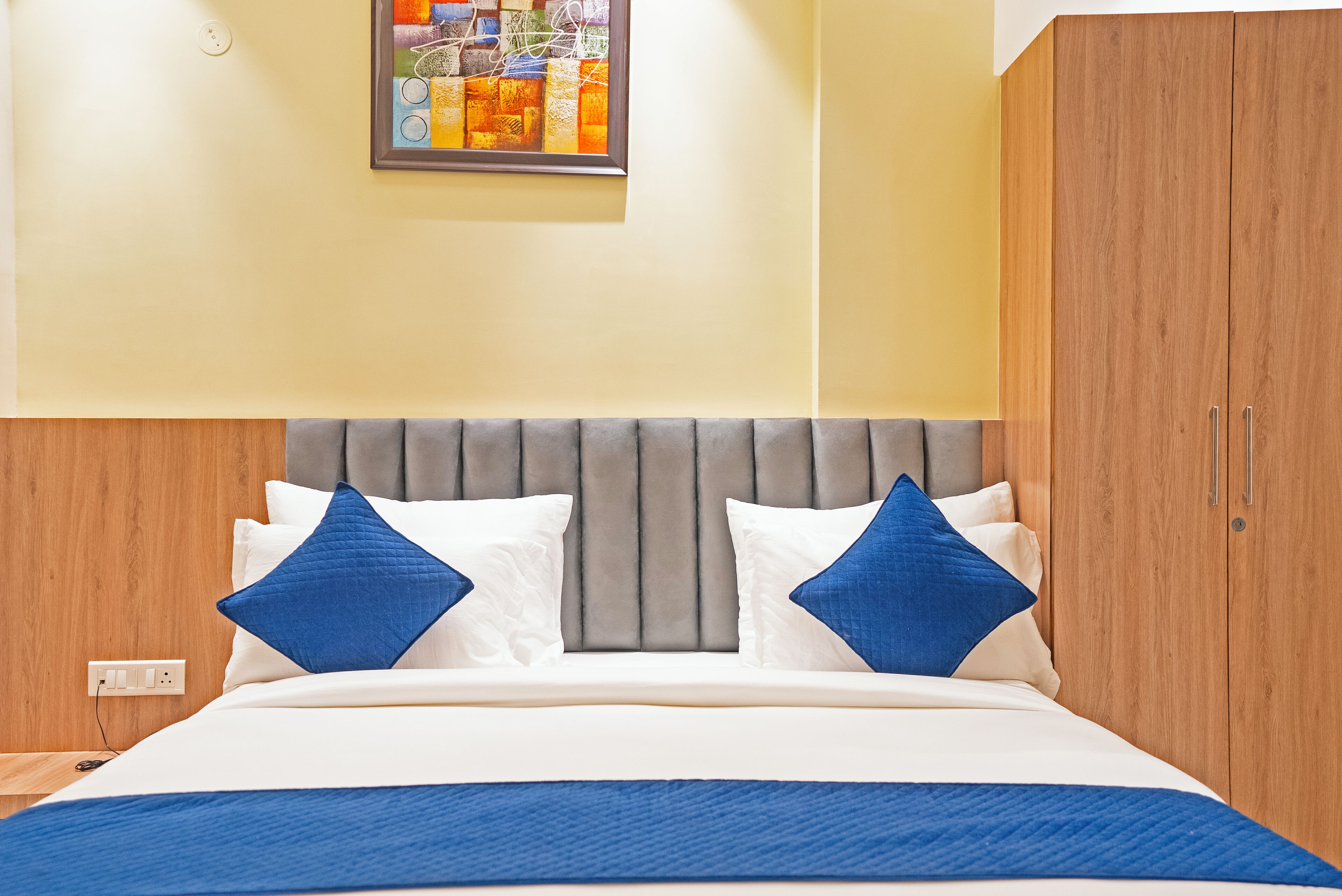 A neatly made bed with a grey upholstered headboard, white and blue pillows, and a blue throw blanket. A wooden wardrobe is to the right, and a colorful abstract painting hangs on the wall above the bed.