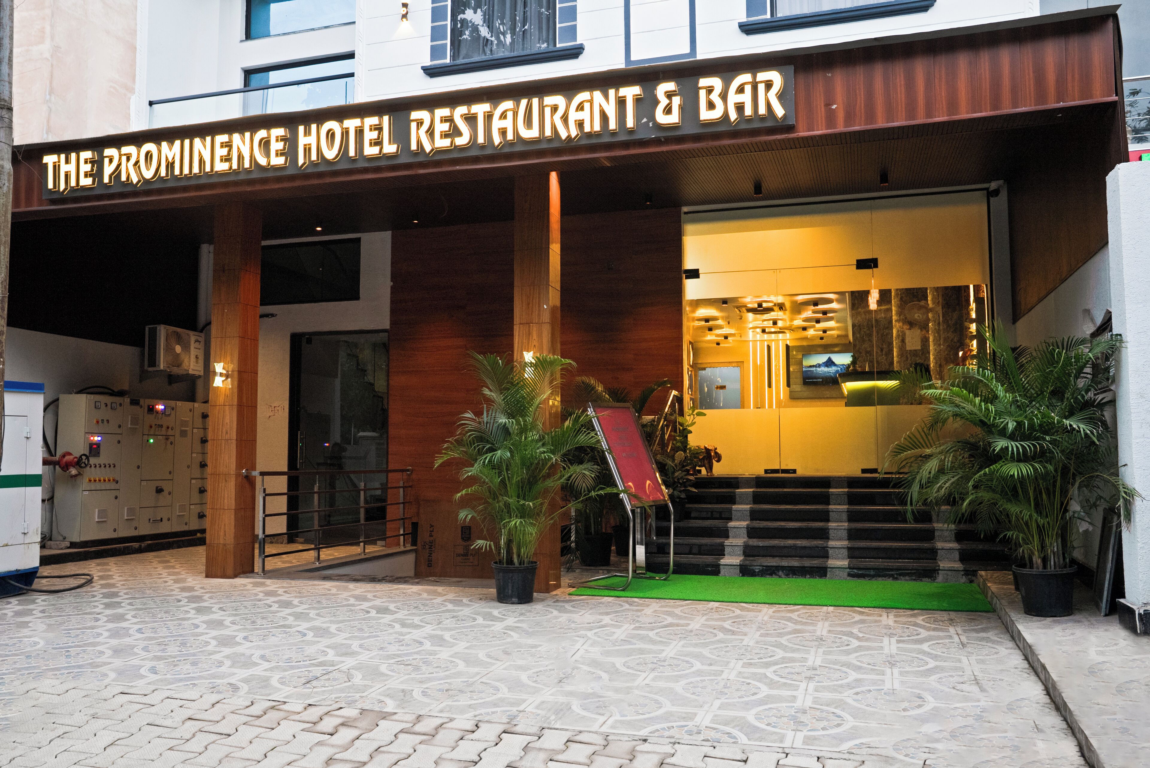 The exterior of The Prominence Hotel Restaurant & Bar with a wooden facade, a green carpeted entrance, and potted plants.