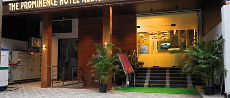 The exterior of The Prominence Hotel Restaurant & Bar with a wooden facade, a green carpeted entrance, and potted plants.