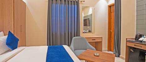 A hotel room with a bed, a desk, a chair, a small table, an air conditioning unit, a mirror, and a door.