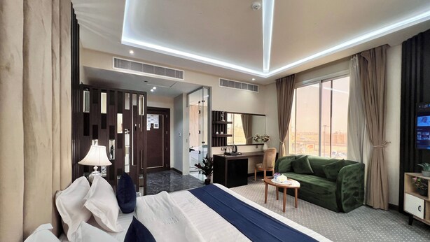 A hotel room with a bed, a desk, a green sofa, a TV, and a large window with curtains. - AL Ewan Pro Serviced Apartments (Al Kharj)