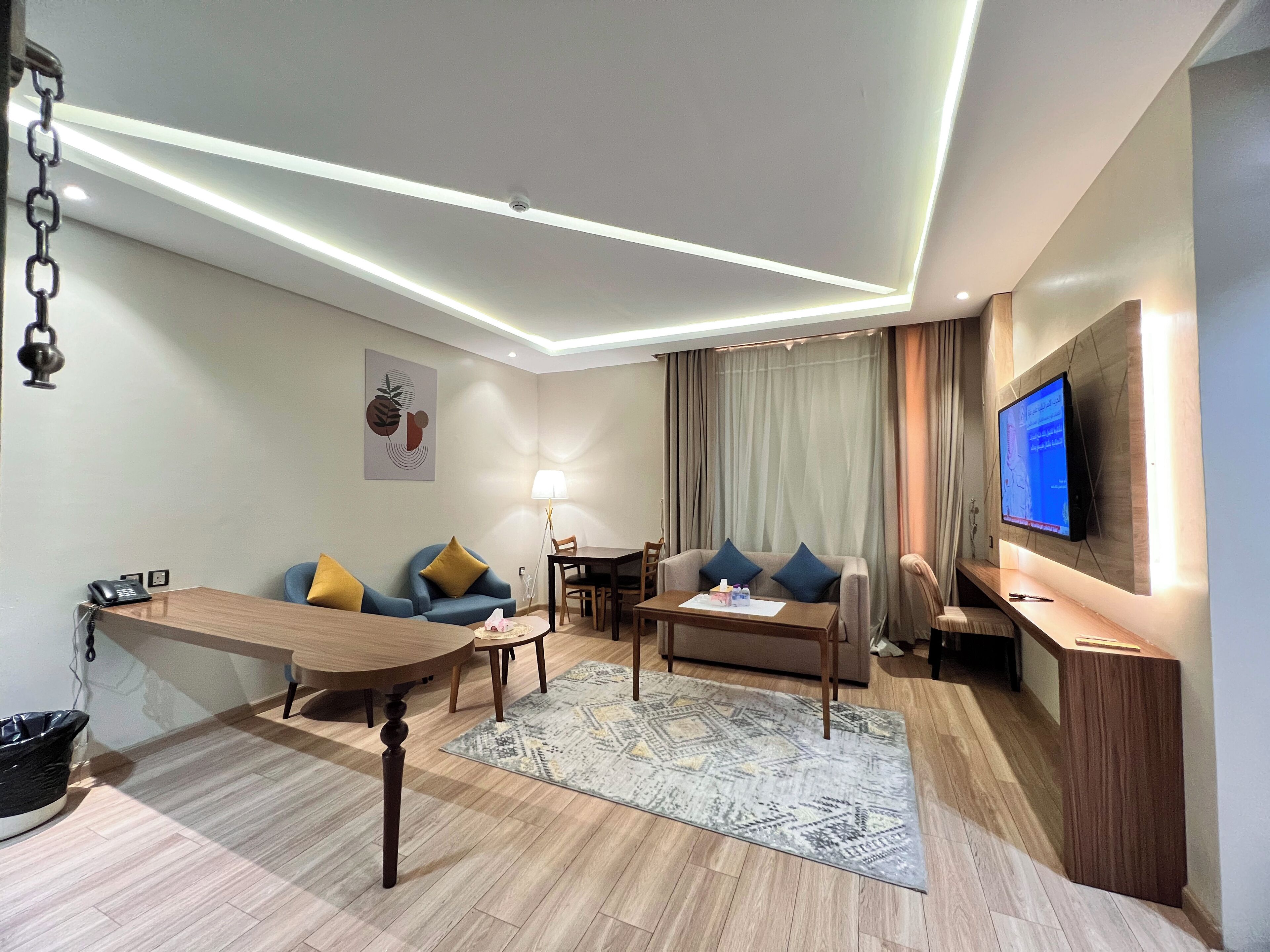 A modern living room with a round wooden table, two sofas with cushions, a patterned rug, a TV on the wall, and a ceiling light fixture.