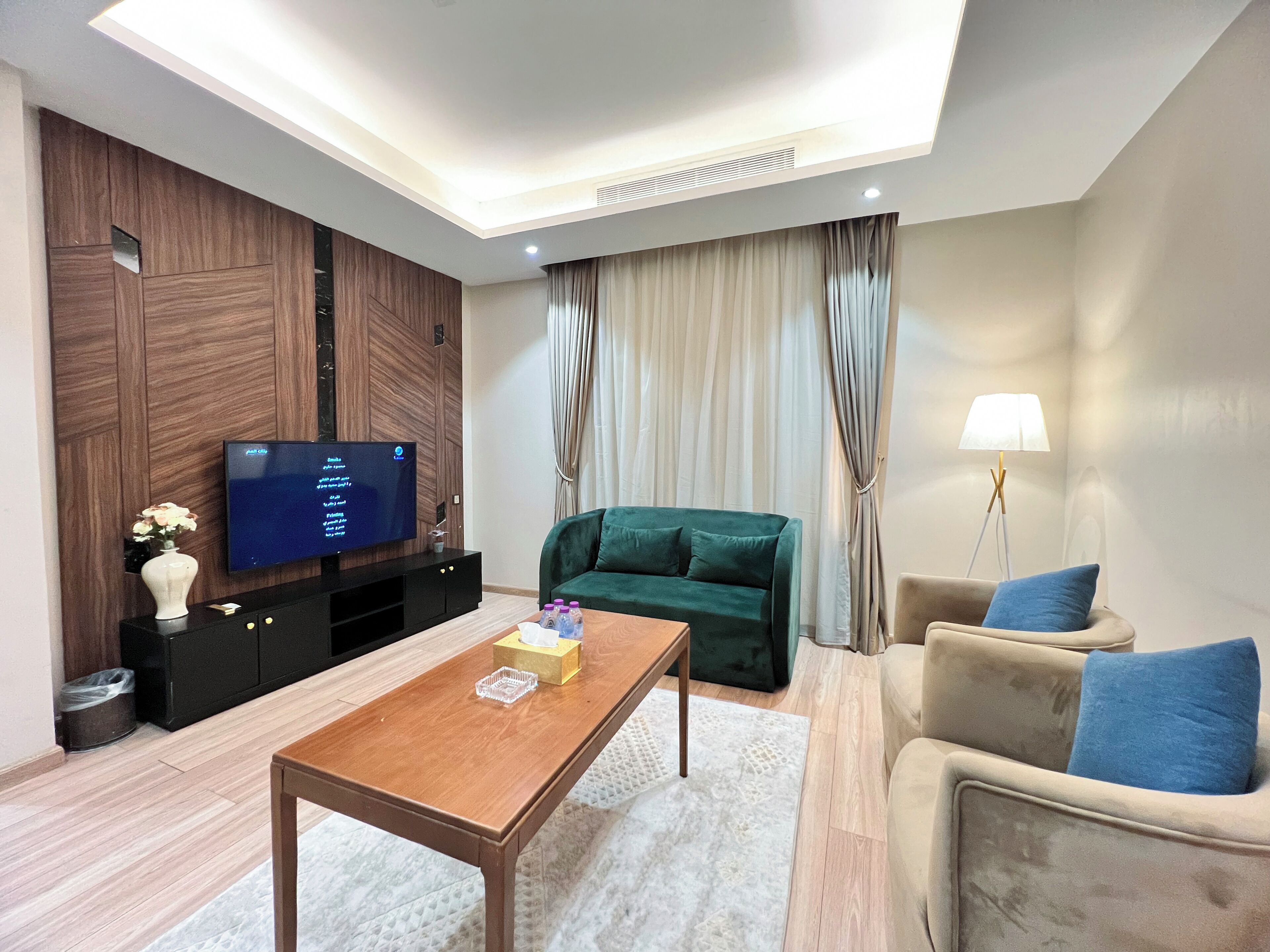 A modern living room with a flat-screen TV, a green sofa, a beige armchair, and a wooden coffee table.