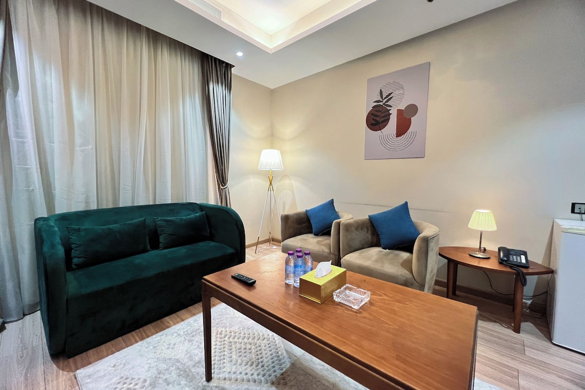 A modern hotel room with a green sofa, a beige sofa with blue cushions, a wooden coffee table, a small side table with a lamp, and a refrigerator.