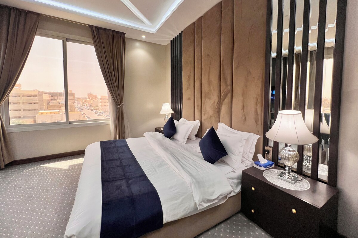 A modern hotel room with a large bed, two bedside tables, a nightstand, a window with curtains, and a city view.