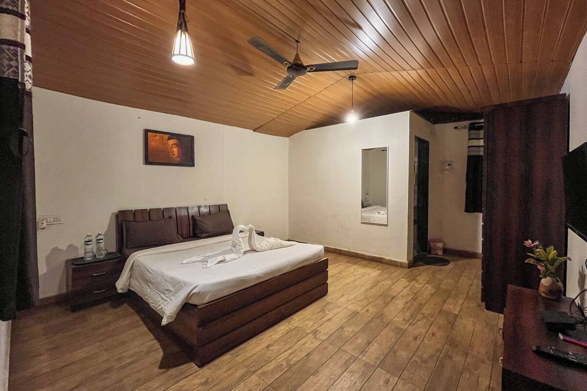 A bedroom with a wooden ceiling, a bed with a headboard, a bedside table with water bottles, a mirror, and a TV on a stand.