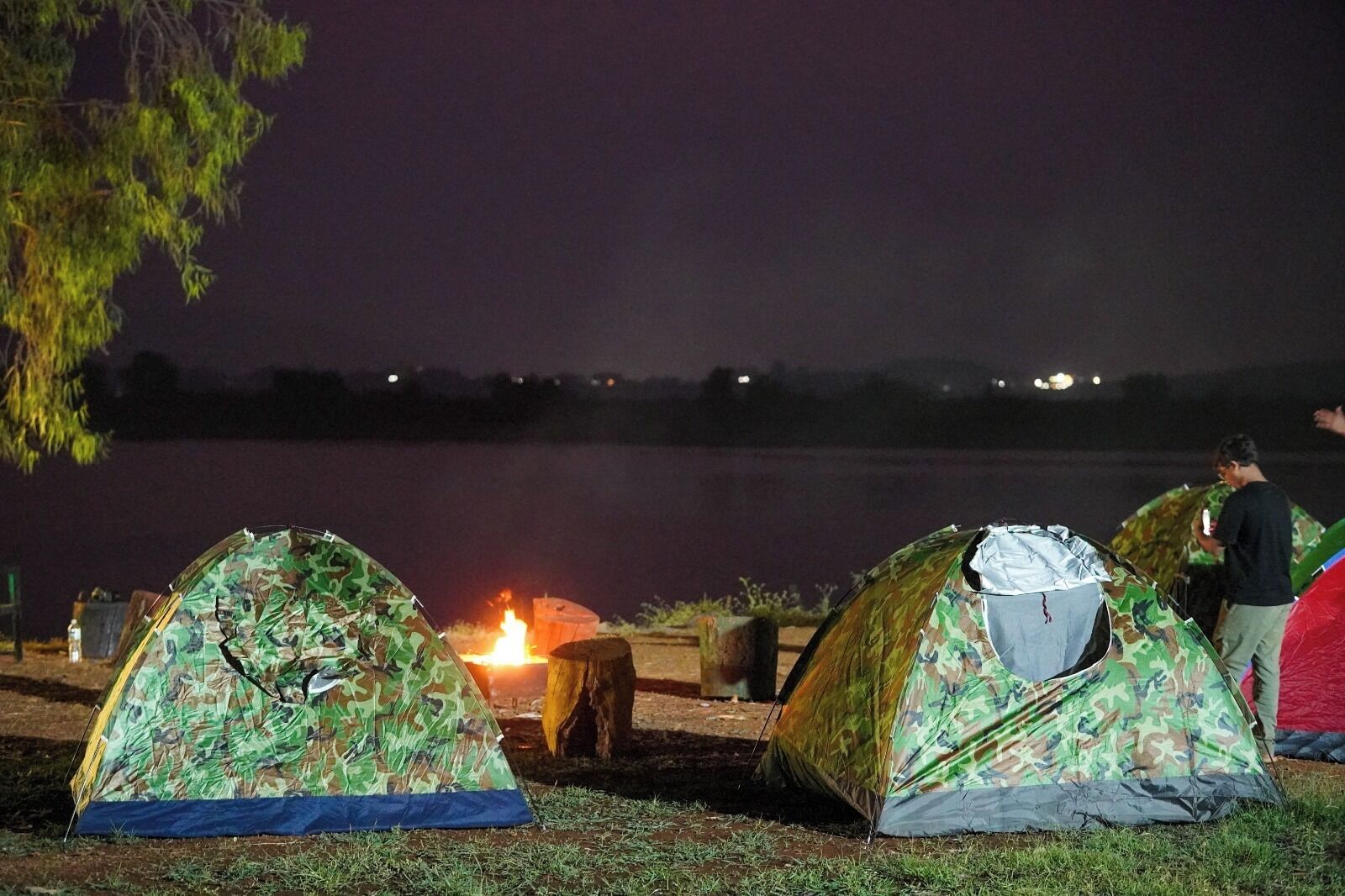 Two camouflaged tents set up near a lake with a campfire between them.