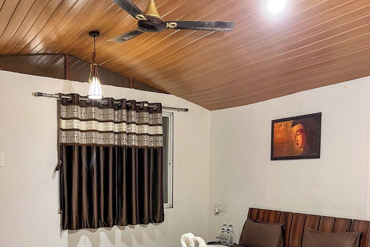 A hotel room with a wooden ceiling, a bed with a white duvet, a ceiling fan, a wall-mounted light, a Buddha painting, and a window with patterned curtains.
