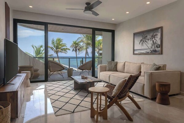 A modern living room with a large sliding glass door leading to a balcony with a view of palm trees and the ocean.