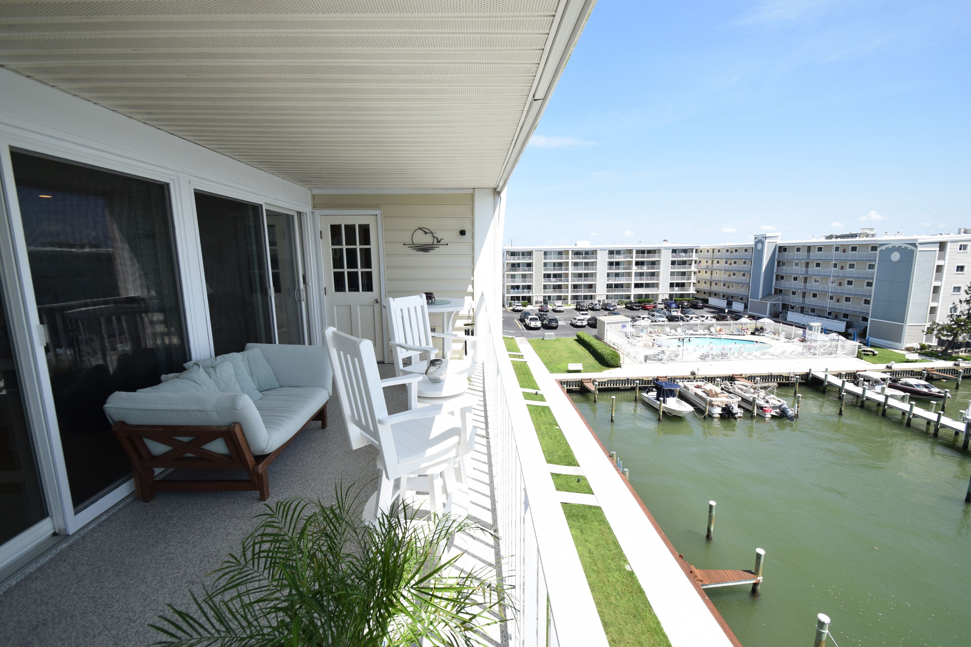 A balcony with a white sofa and chairs, overlooking a marina with boats and a pool.