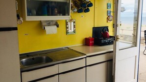 A kitchen with a yellow wall, a sink, a stove, and a window overlooking a beach. - Comfy Holiday Home & Beach Hut (Sutton on Sea)