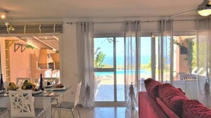 A modern living room with a red sofa, dining area with chairs and a table, and a large sliding glass door leading to a pool and ocean view. - Exclusive Golf Villa (Private Swimming Pool) Costa Adeje (Adeje)