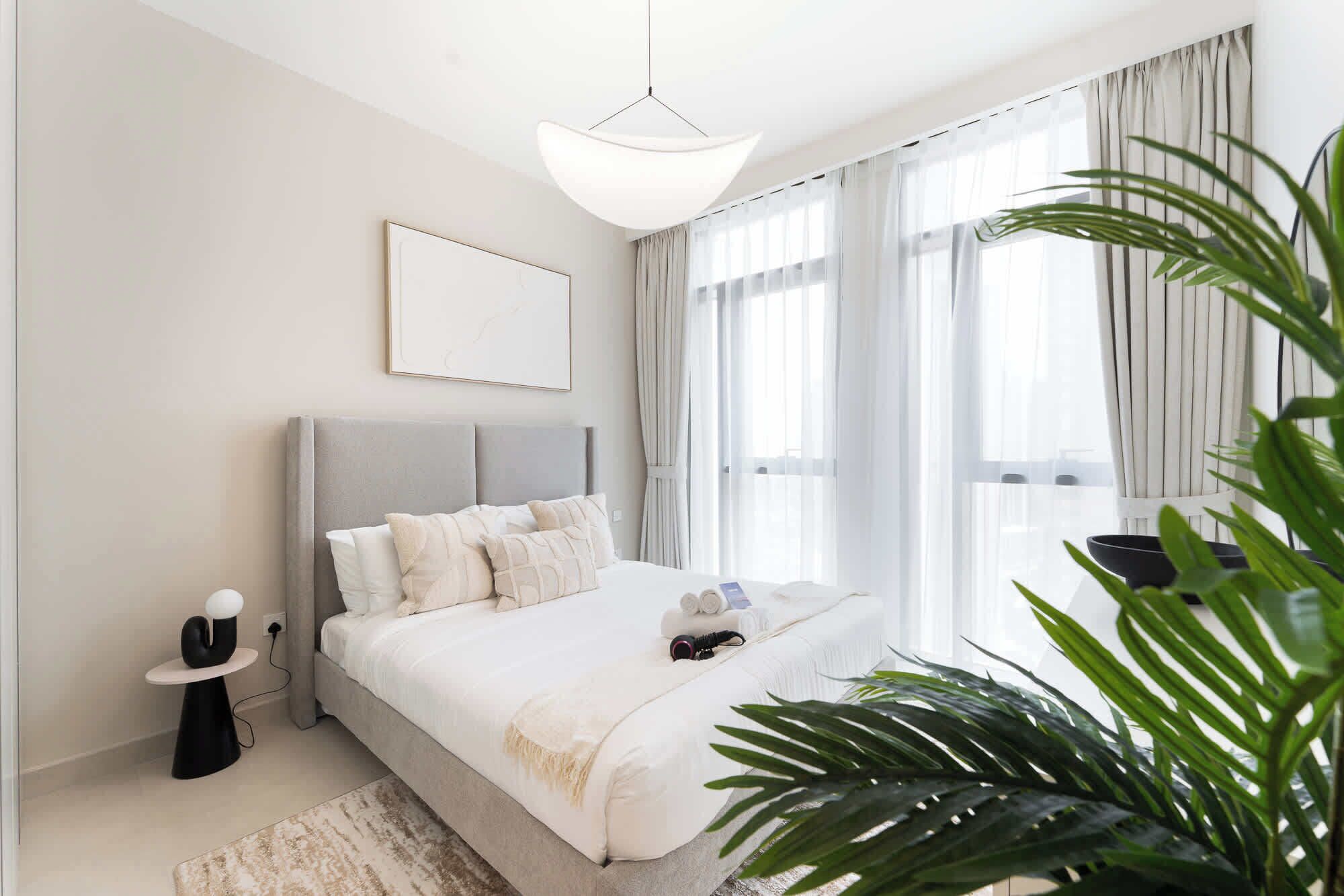 A modern bedroom with a large bed, a bedside table, a hanging light fixture, and a window with sheer curtains.