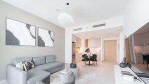 A modern living room with a grey sofa, a coffee table, and a rug. There is a dining area with a table and chairs. The kitchen is visible in the background with cabinets and appliances.