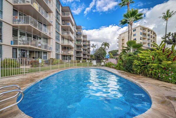 A swimming pool surrounded by multi-story buildings with balconies, palm trees, and a clear sky.